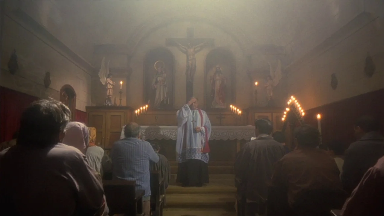 Dagon (2001) movie still: wide — A small dim chapel features a wooden altar with a central crucifix and statues. The priest stands before the altar,…