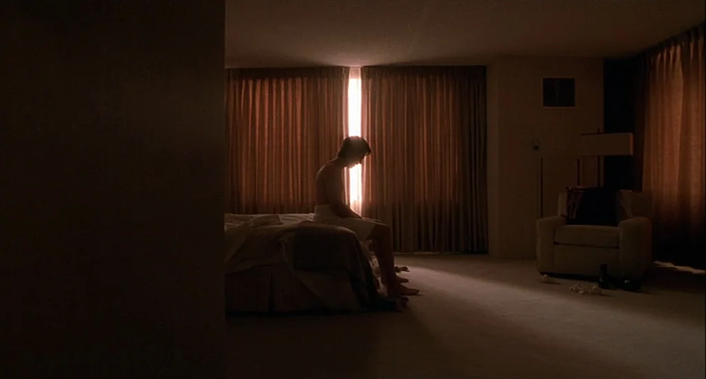 One Hour Photo (2002) movie still: wide — A spacious hotel room features beige carpet, brown curtains, and a single upholstered chair. A man sits…