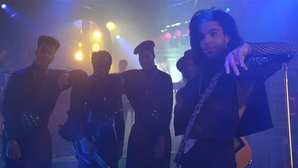 Graffiti Bridge (1990) movie still: medium — A dimly lit musical performance area on a concert stage with hazy atmosphere. Prince stands in the…