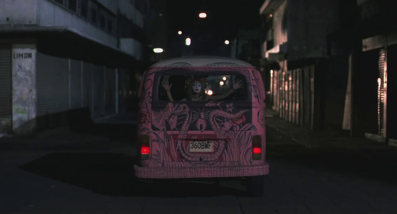Santa Sangre (1989) movie still: wide — A dark, narrow urban street at night with a parked pink decorated van. A woman with white face makeup peers…