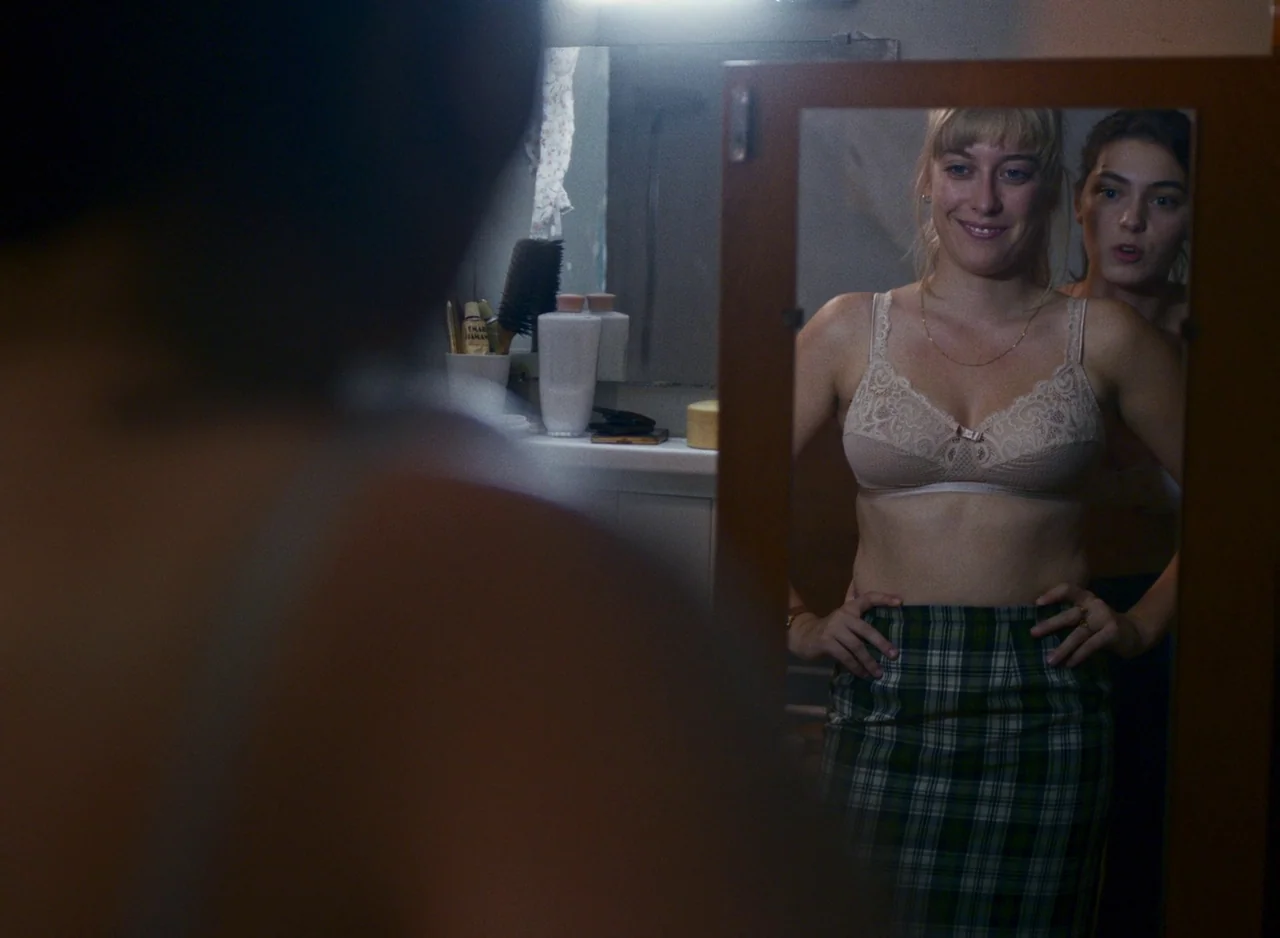Happening (2021) movie still: medium — A dimly lit room with a wooden-framed mirror hanging on a wall. Two women stand side-by-side looking into the…