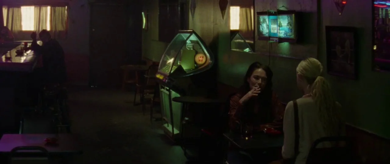 Low Down (2014) movie still: wide — A dimly lit, sparse dive bar features green-painted walls and a vintage jukebox interior. Two women sit at a…