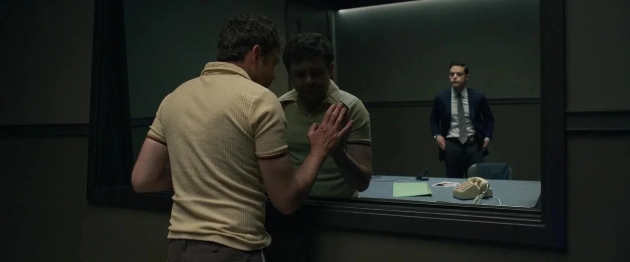 The Little Things (2021) movie still: medium — A man stands before a reflective glass partition in a stark, dimly lit room. Behind the glass, a man…