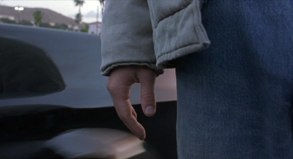 Brick (2005) movie still: closeup — A dark, reflective car surface sits in a parking lot during the dusk. A person wearing a tan puffer jacket and…