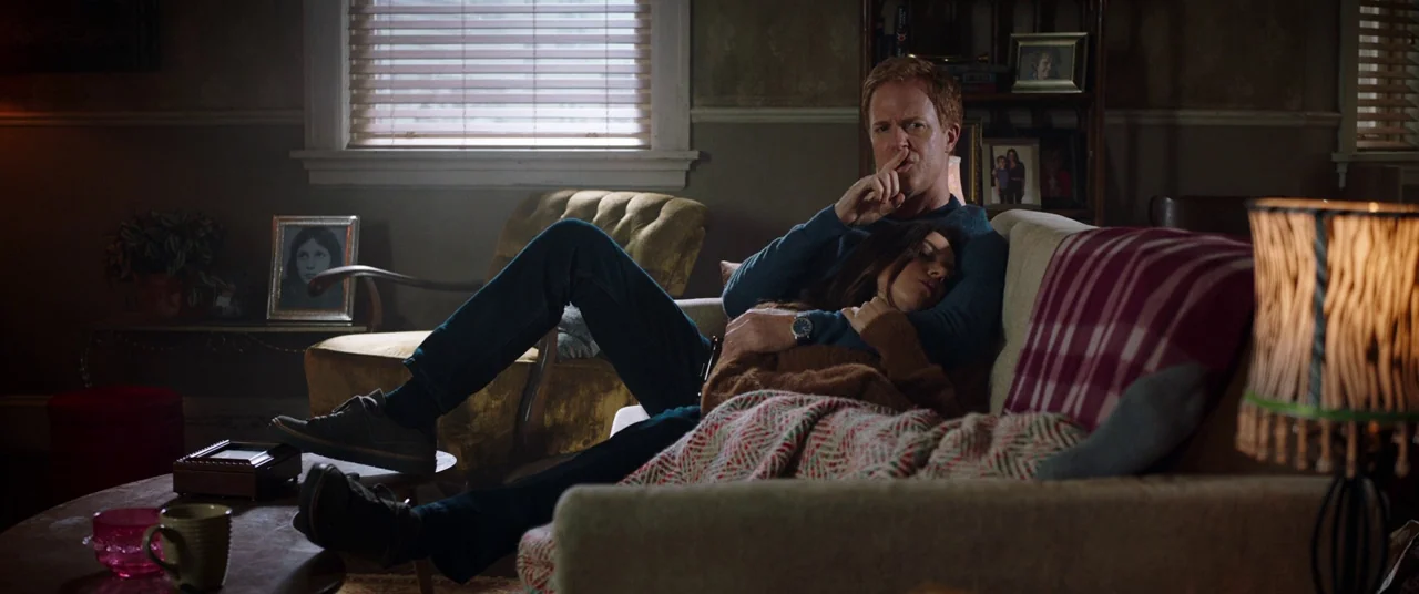 Child's Play (2019) movie still: medium — A dimly lit room with closed window blinds and personal framed photos on furniture. A man sits on a couch…