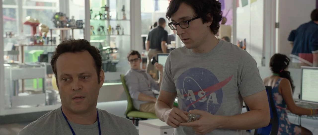 The Internship (2013) movie still: medium — A modern open-plan office features glass walls and shelves filled with various toys. Two men are engaged…