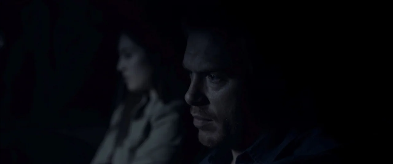 Radius (2017) movie still: medium — A dark vehicle interior with a man in the foreground and a woman behind. The man looks forward while the woman…
