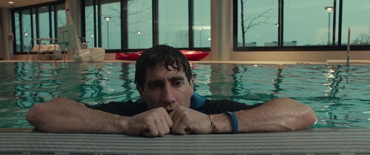 Stronger (2017) movie still: medium — A modern indoor swimming pool area features glass walls and a gray tiled deck. A man with wet hair rests his…