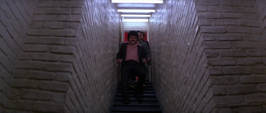 Carlito’s Way (1993) movie still: medium — A narrow stairwell with white painted brick walls and metal stairs. A group of men descend the stairs in…