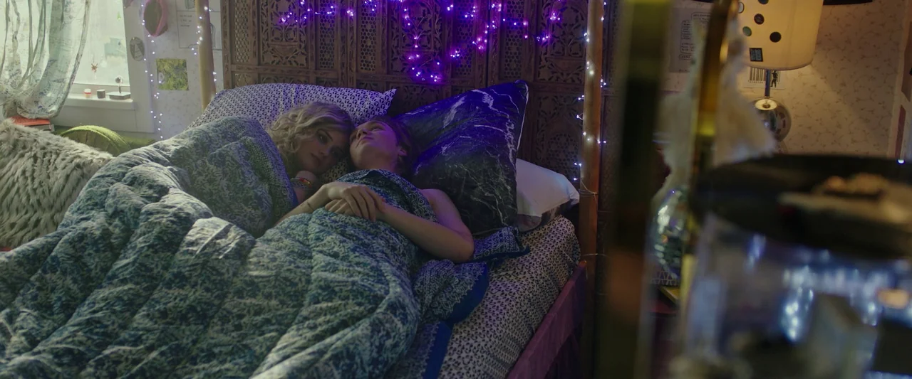 Spontaneous (2020) movie still: medium — A shared bed features a large carved wooden headboard adorned with purple fairy lights. Two young people…