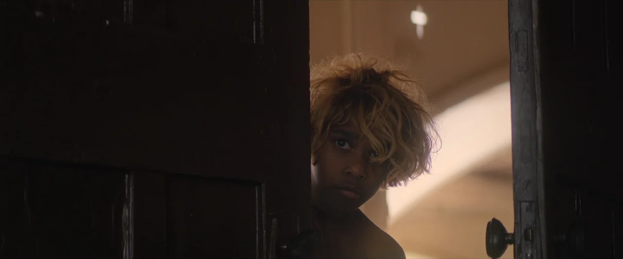 The New Boy (2023) movie still: closeup — A dark wooden door is partially ajar, creating a narrow opening into a room. A young person with messy…