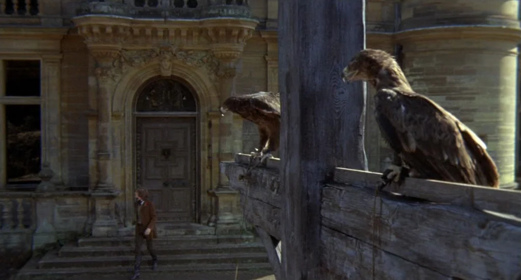 Demons of the Mind (1972) movie still: wide — A stone mansion facade features a large arched door and intricate architectural carvings. Two hawks…