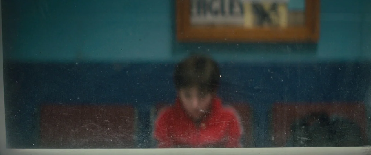 Brightburn (2019) movie still: medium — A person in a red hoodie sits on a bench behind a smudged window. A blue wall and a framed picture are…