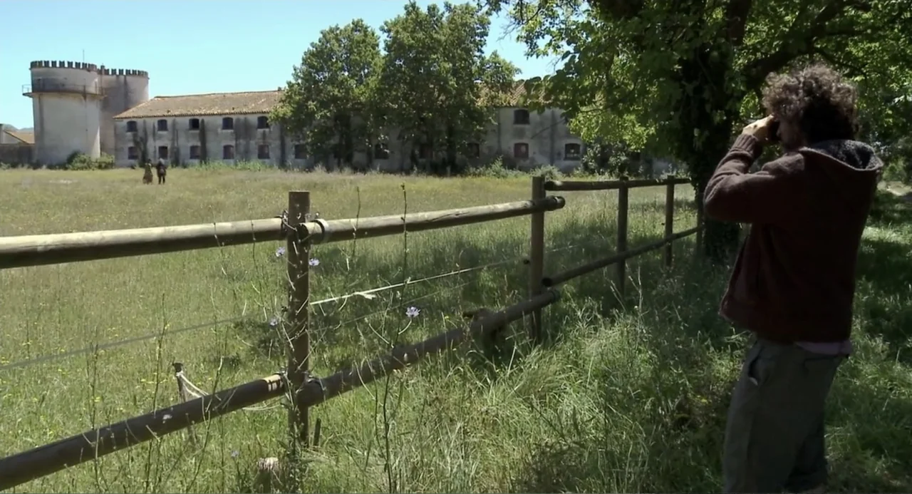 Az X- Kryptonit projekt (2020) movie still: medium — A sunlit grassy field with a wooden post fence sits before a distant building. A man in a brown…