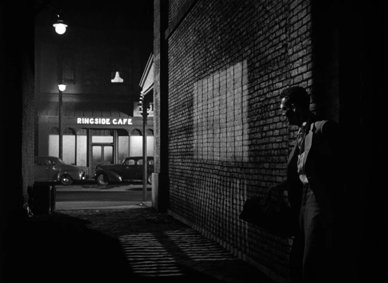 The Set-Up (1949) movie still: medium — A dark city alleyway at night featuring a large brick wall with shadow projections. A man in a suit walks…