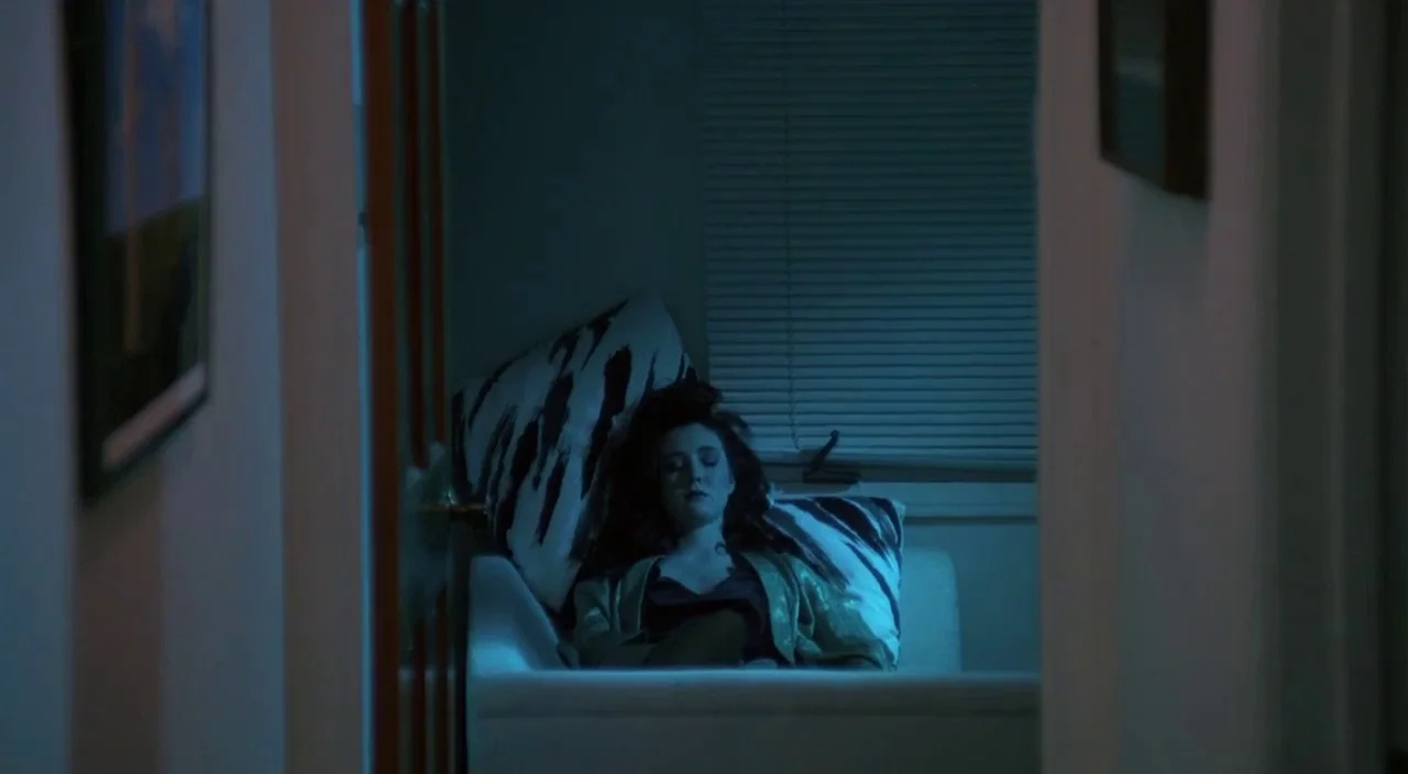 Heathers movie still: medium — A dimly lit room with blue light filtering through horizontal blinds on a window. A woman with dark hair leans back…