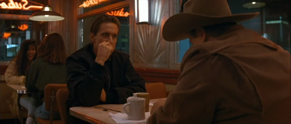 Basic Instinct (1992) movie still: medium — A dark diner booth at night with amber neon signs visible through the windows. Two men sit at a table…
