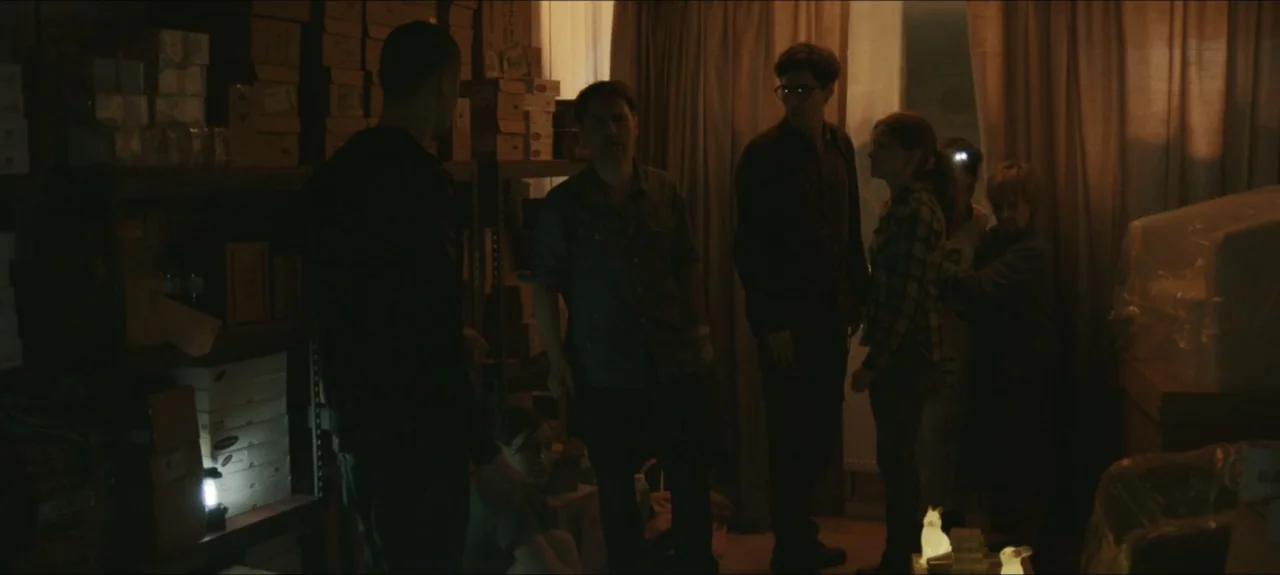 Containment (2015) movie still: medium — A cluttered storage room filled with stacked cardboard boxes on metal shelving units. A group of five…