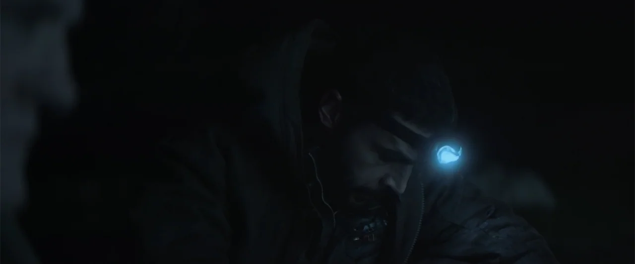 Bring Them Down (2025) movie still: closeup — A man wearing a headlamp stands in a dark, outdoor nocturnal environment. He leans forward with his…