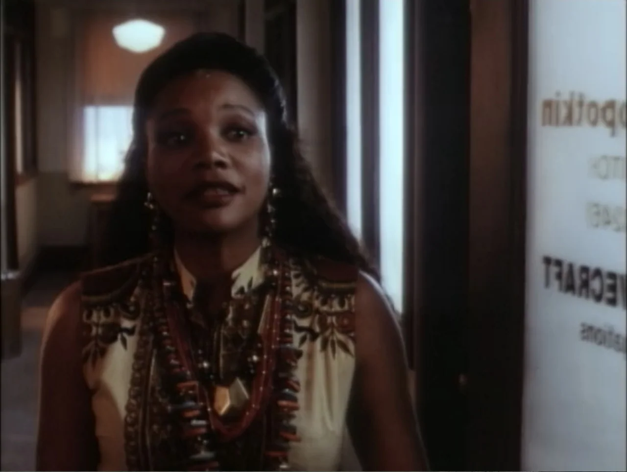 Cast a Deadly Spell (1991) movie still: medium — A dimly lit indoor hallway with a glass door displaying reversed text labels. A woman in a…