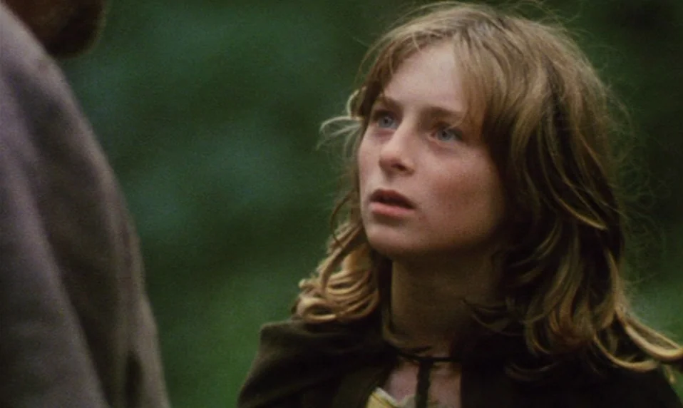 Black Jack (1979) movie still: closeup — A dense forest environment with a soft green backdrop of out-of-focus foliage. A young woman with wavy hair…