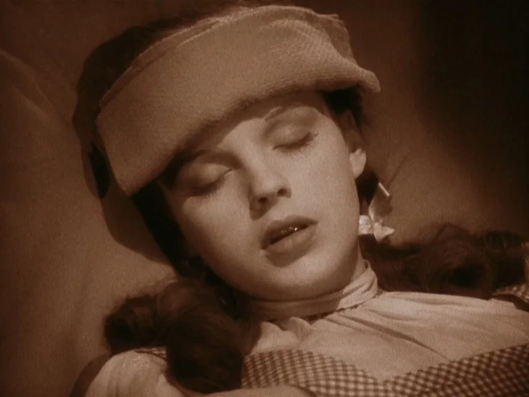 The Wizard Of Oz (1939) movie still: closeup — A person lies on a soft surface with a cloth folded across their forehead. The subject sleeps soundly…