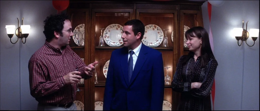 Punch Drunk Love (2002) movie still: medium — A formal interior space features a wooden china cabinet displaying decorative plates and glasses. Two…