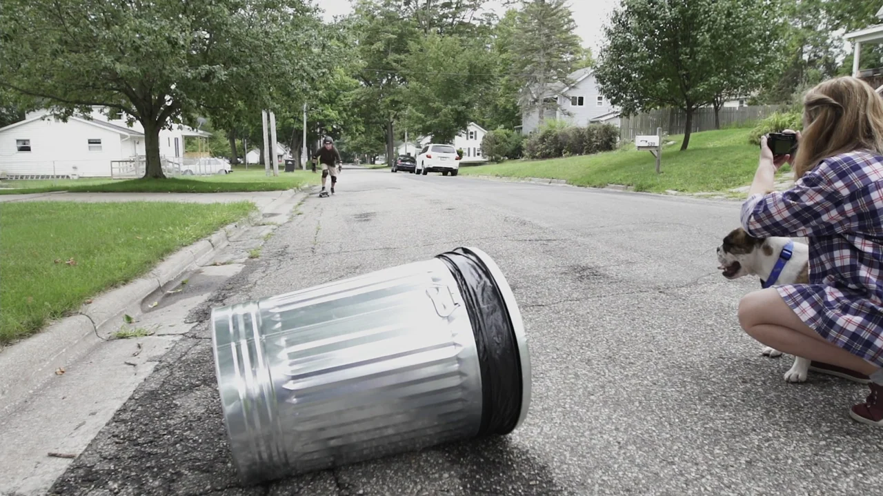 Sk8 Dawg (2018) movie still: medium — A quiet suburban street with houses and trees features a metal trash can foreground. A woman kneels with a dog…