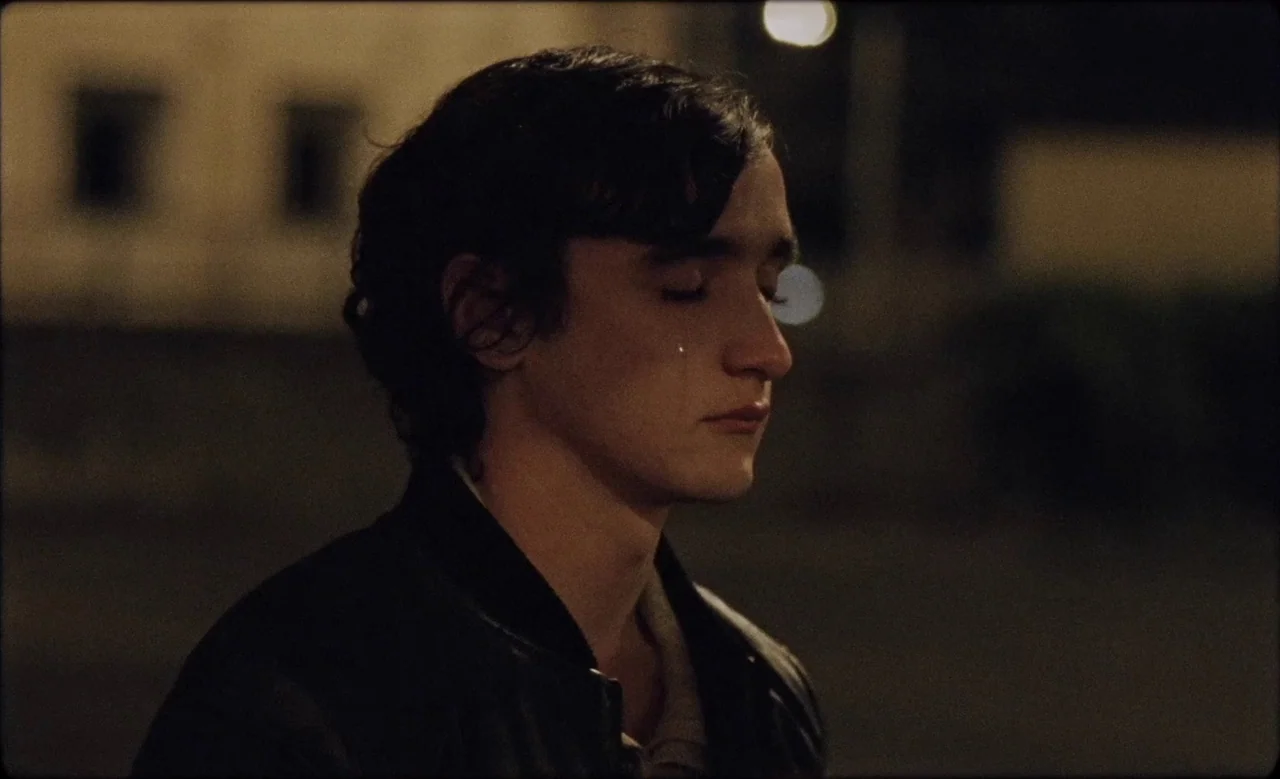 Happy as Lazzaro (2018) movie still: closeup — A dark outdoor urban setting illuminated faintly by a blurred background streetlamp. A young man in a…