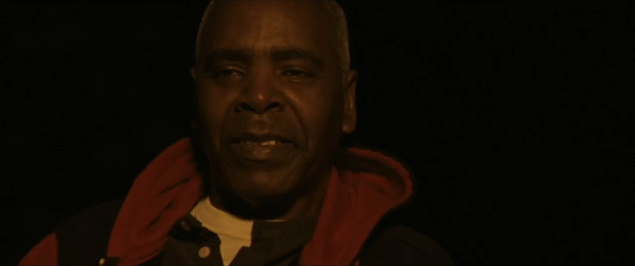 Sermon on the Mount (2019) movie still: closeup — The subject stands in an entirely dark, unidentified indoor space. A man wearing a red-hooded…