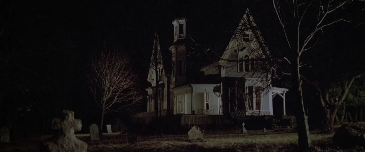 House By The Cemetery (1981) movie still: establishing — A gothic-style house sits in a dark, empty field filled with old stone graves. The…
