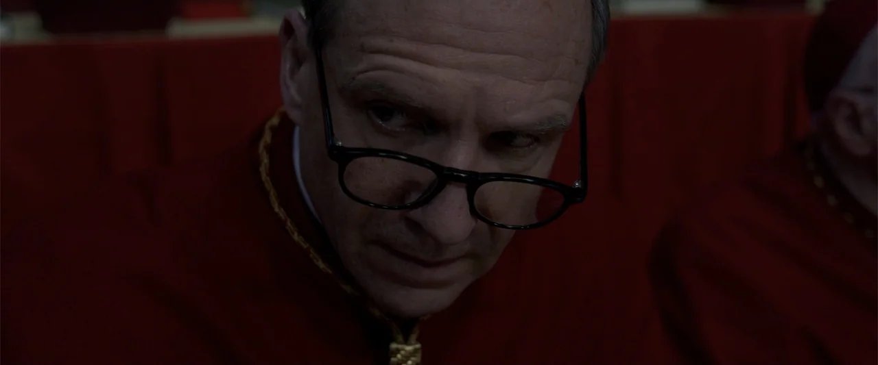 Conclave (2024) movie still: closeup — A man in clerical robes sits within a dimly lit crimson chamber. The subject wears dark rimmed glasses and…