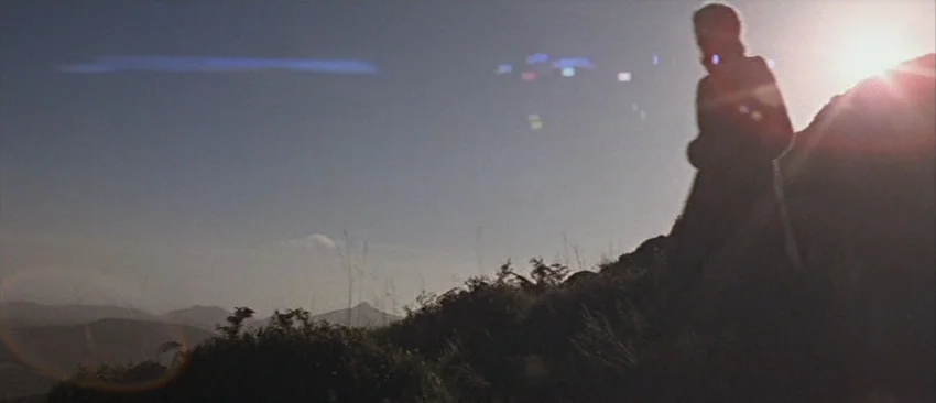 Images (1972) movie still: fullbody — A grassy hillside slopes upward under a clear sky during the day. A man in an overcoat walks up the incline…