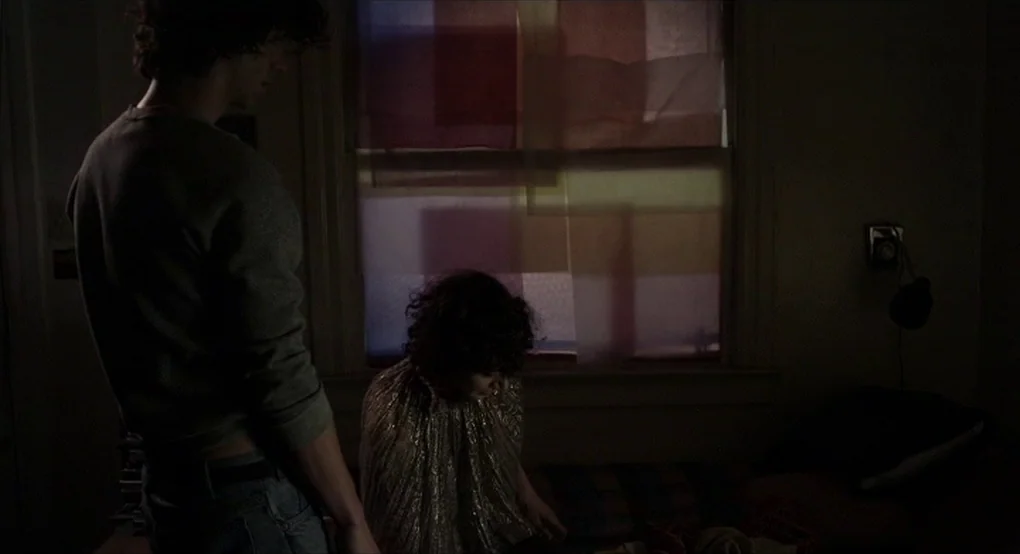 The Future (2011) movie still: medium — A dark, dimly lit space features a window covered in multicolored translucent paper squares. A man stands…