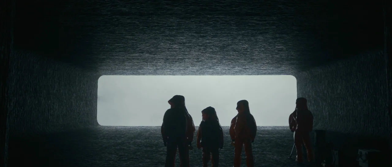 Arrival (2016) movie still: wide — A dark, textured tunnel with a rectangular opening revealing a bright, overcast exterior sky. Four figures in…