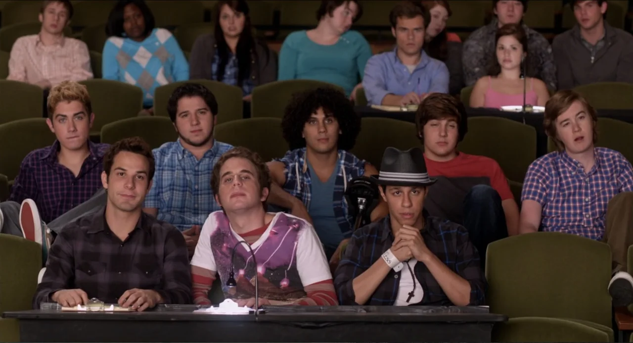 Pitch Perfect (2012) movie still: medium — A tiered lecture hall features rows of green padded seats filled with young adults. Three young men sit…