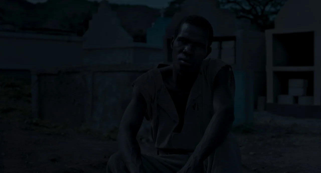 Zombi Child (2019) movie still: medium — A dark, dimly lit cemetery features multiple concrete grave structures in the background. A young man sits…
