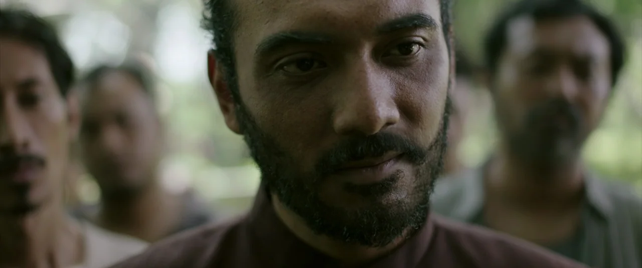 Impetigore (2019) movie still: closeup — A group of men stand together outdoors in a blurry, natural environment. The primary subject in the…