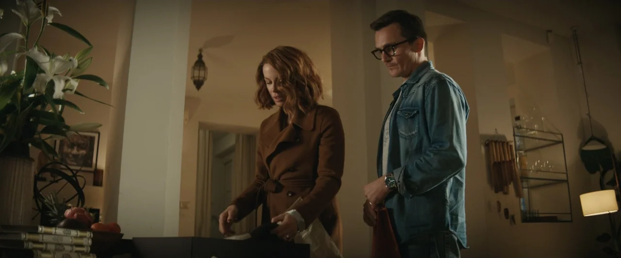 Canary Black (2024) movie still: medium — An interior space features a table with a white flower vase and various decor items. A woman in a brown…