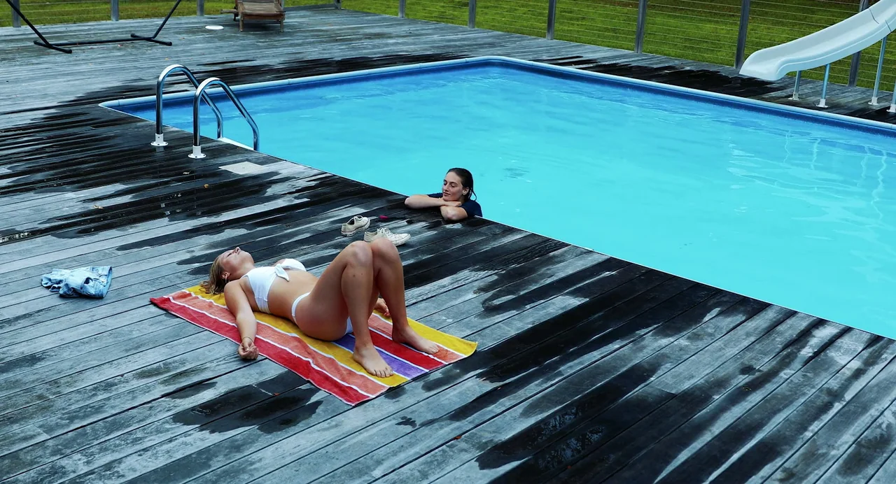 Hellbender (2021) movie still: wide — A wooden deck surrounds a bright blue pool with a white plastic slide. One woman lies on a colorful striped…
