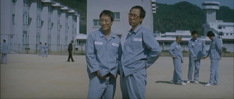 Mother (2009) movie still: fullbody — A spacious, gravel-covered yard surrounded by multi-story institutional grey buildings. Multiple male figures…