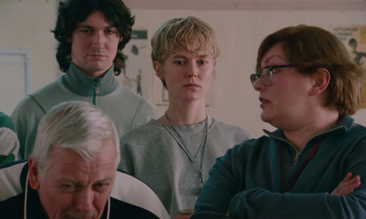 Blue Jean (2022) movie still: medium — A group of four people gather inside an indoor athletic or recreational facility. A woman wearing glasses…