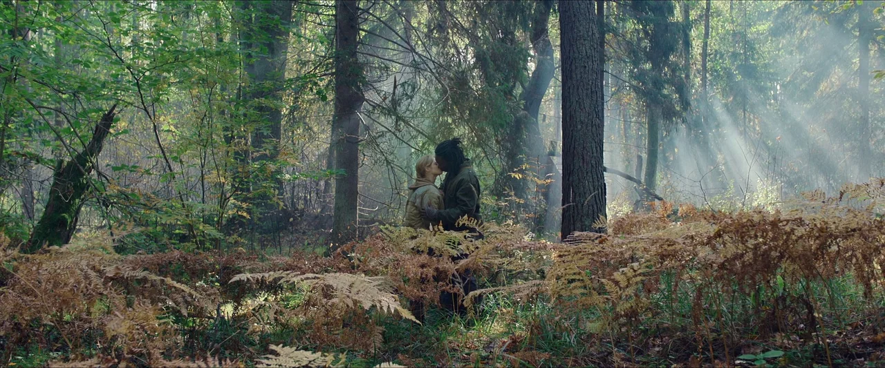 Azrael (2024) movie still: wide — A dense forest with tall pine trees and thick undergrowth of dried ferns. A man and a woman stand together in the…