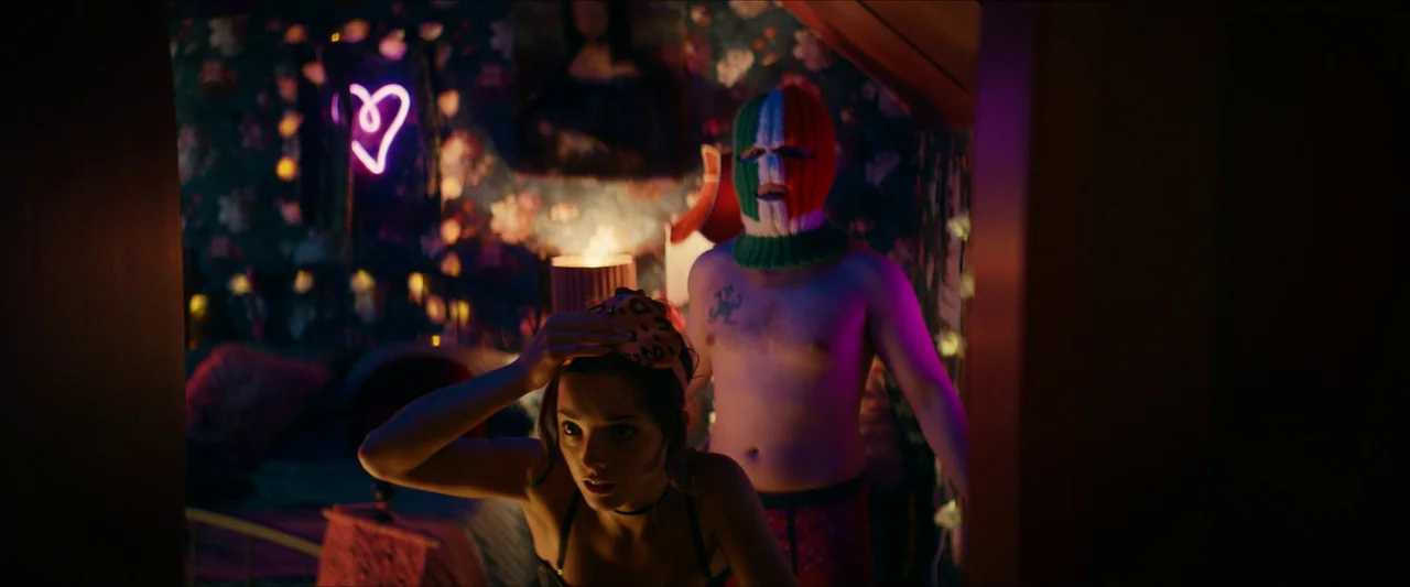 Kneecap (2024) movie still: medium — A cluttered room features dark walls decorated with floral wallpaper and a neon heart sign. A woman lifts an…