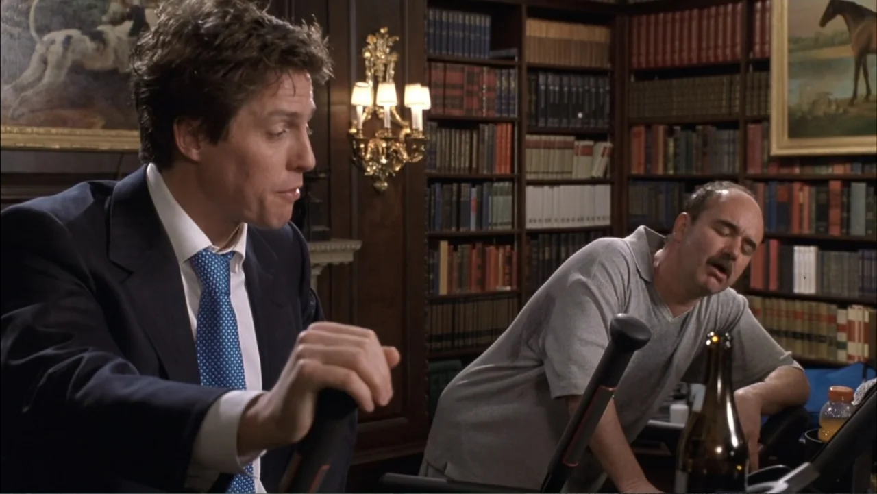Two Weeks Notice (2002) movie still: medium — A wood-paneled office contains tall bookshelves filled with books and an exercise bike in front. Two…