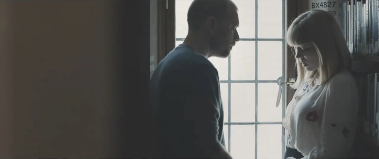 Zoe (2018) movie still: medium — A quiet corner of a room features a tall bookshelf next to a grid-paned window. A man stands profile-to-camera…