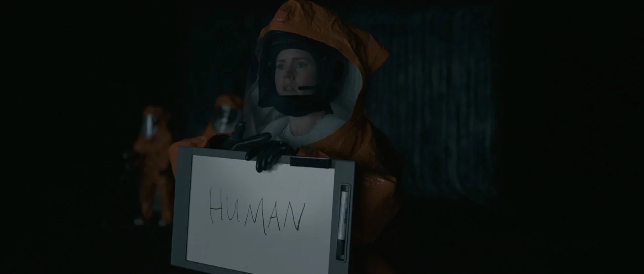 Arrival (2016) movie still: medium — A dark, cavernous interior with non-human architecture and deep shadows. A woman in an orange hazmat suit holds…