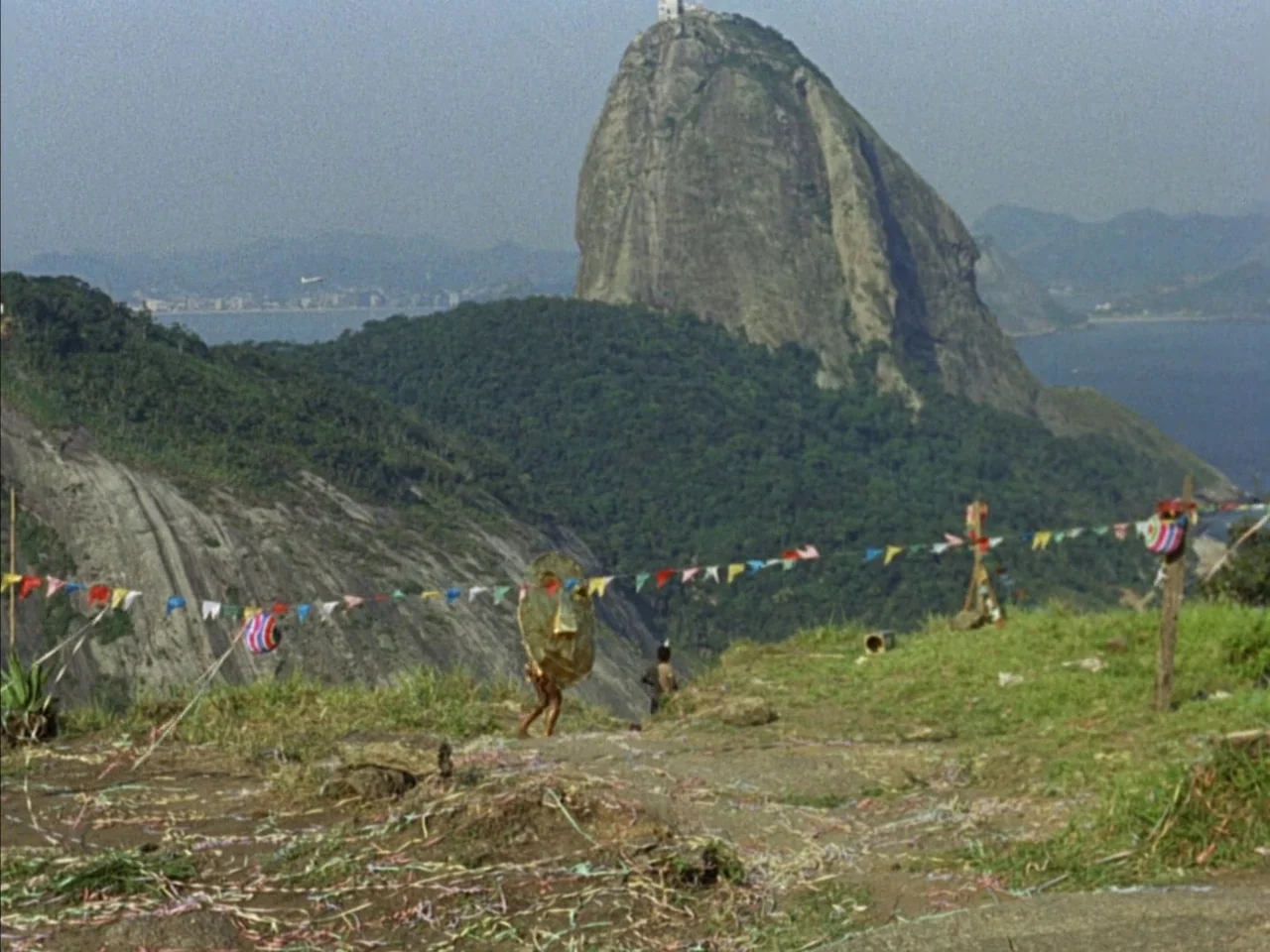 Black Orpheus (1959) movie still: wide — A grassy hillside overlook features a large mountain peak in the distant background. A person walks across…