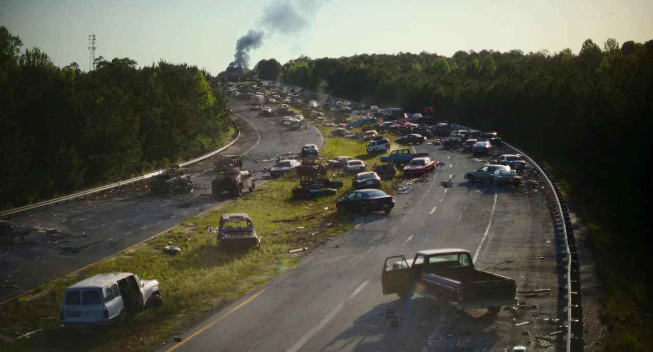 Civil War (2024) movie still: establishing — A long stretch of highway is littered with dozens of abandoned and damaged vehicles. Dark smoke rises…