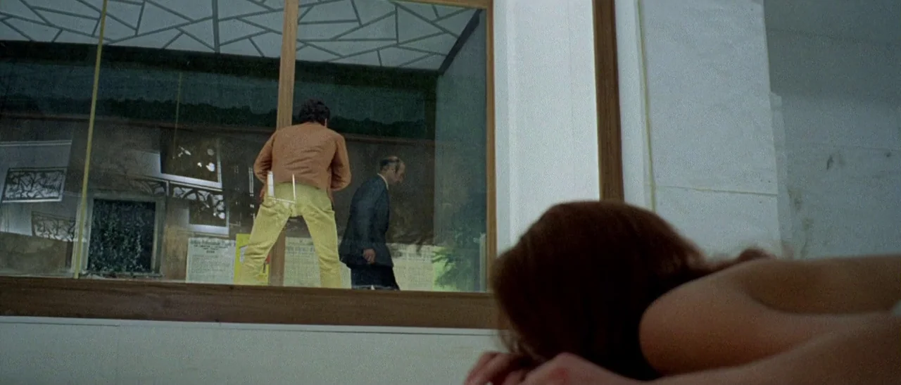 The Bird With The Crystal Plumage (1970) movie still: wide — A modern building with large glass panes stands behind a white wall. Two men walk past…